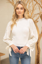 Load image into Gallery viewer, Woman wearing a white blouse with long sleeves and tied cuffs, standing in front of a decorative tree.
