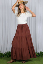 Load image into Gallery viewer, Woman wearing a white top and brown tiered skirt with a hat against a white curtain background.
