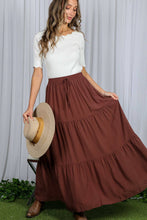 Load image into Gallery viewer, Woman wearing a white top and brown tiered skirt holding a straw hat against a gray curtain background.

