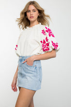 Load image into Gallery viewer, A model wearing a white blouse with pink floral patterns, showcasing the Alice Corduroy Top's cozy comfort and stylish embroidery.
