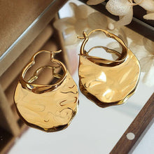 Load image into Gallery viewer, A pair of gold-plated, wavy design hoop earrings on a reflective surface.
